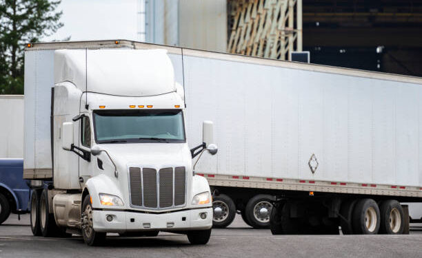 White big rig long haul semi truck with high cab and semi trailer turning on warehouse parking lot waiting for loading and possibility of continuing to the destination according to approved schedule