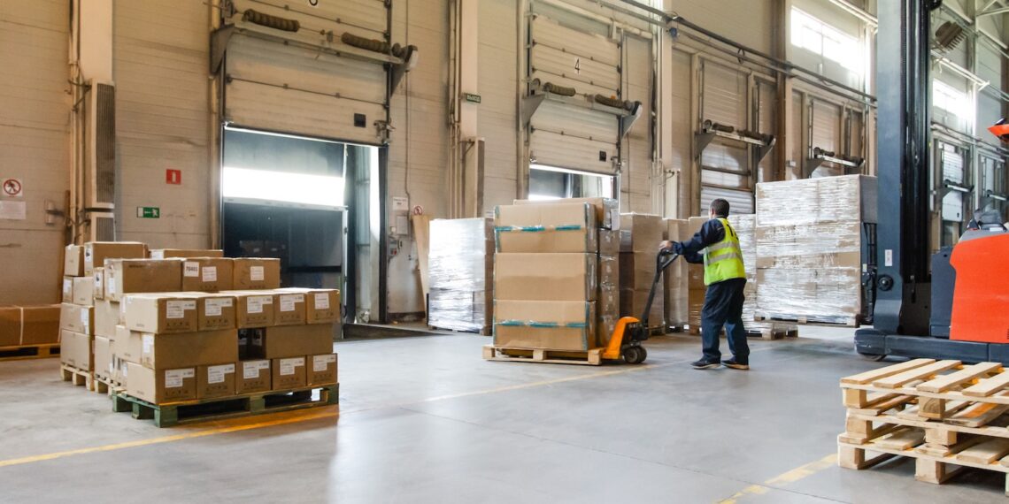 Interior of a modern warehouse storage of retail shop with pallet truck near shelves and loading ramps. Storehouse worker in uniform working on forklift.