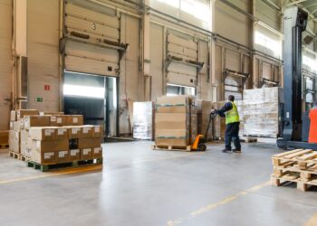 Interior of a modern warehouse storage of retail shop with pallet truck near shelves and loading ramps. Storehouse worker in uniform working on forklift.