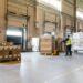 Interior of a modern warehouse storage of retail shop with pallet truck near shelves and loading ramps. Storehouse worker in uniform working on forklift.